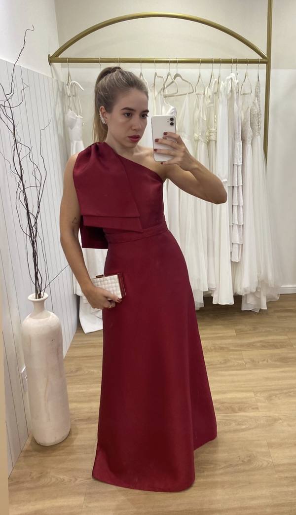 Vestido longo marsala de um ombro só, com laço estruturado no busto e saia evasê de caimento fluido.