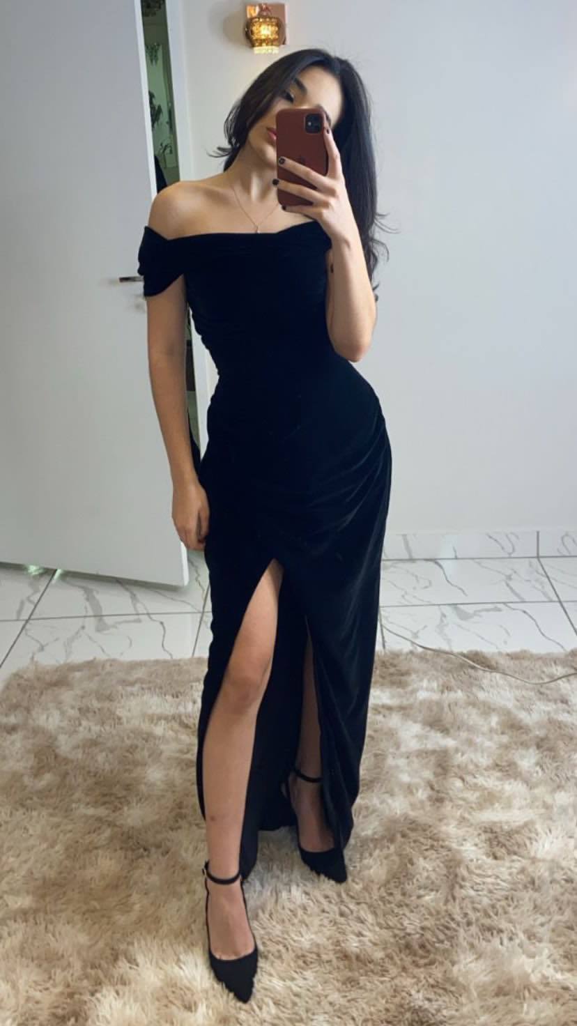 Vestido longo preto em veludo, decote ombro a ombro, caimento ajustado ao corpo e fenda frontal na saia.