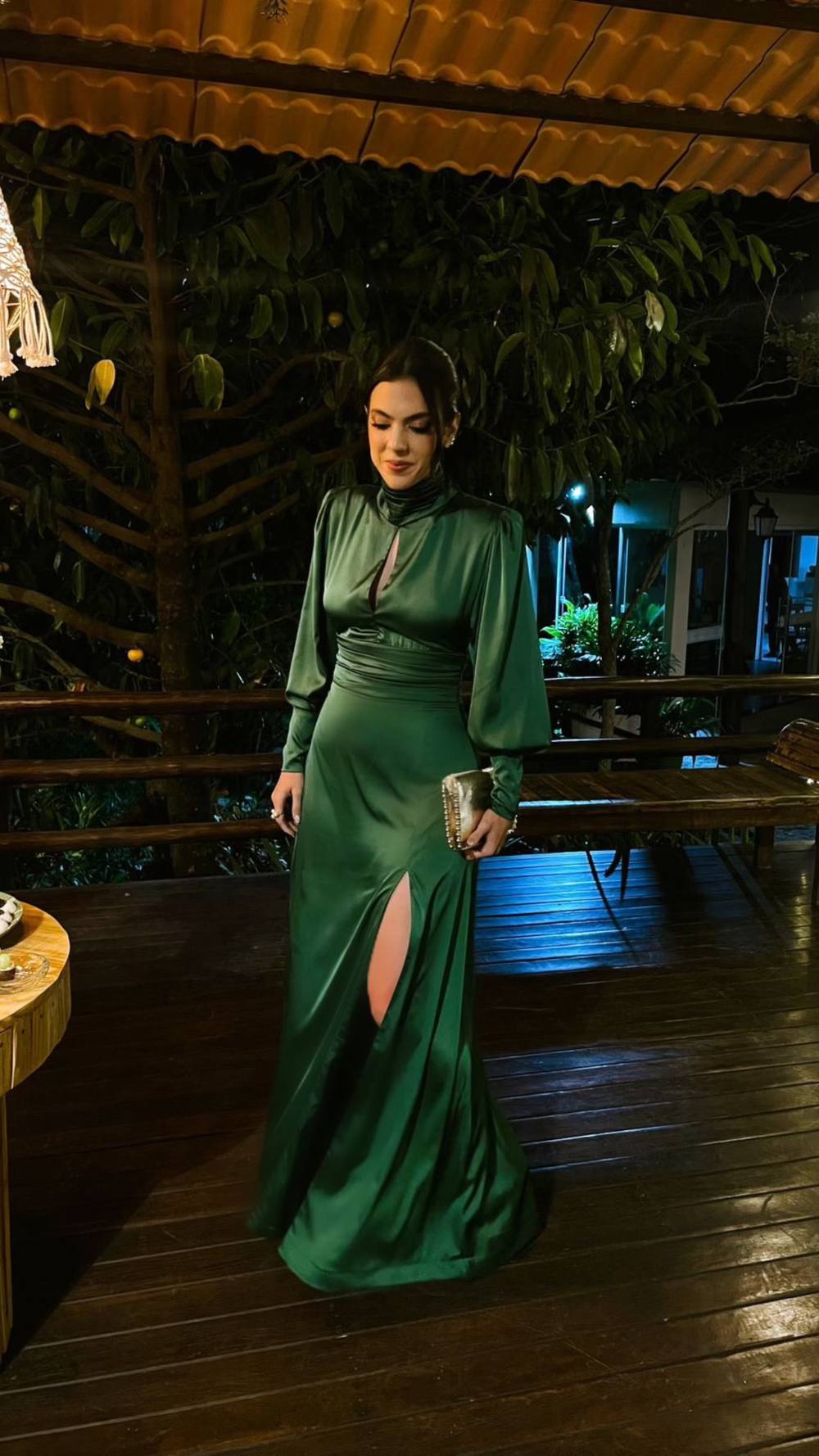 Vestido longo verde de cetim, com manga longa, gola alta, saia fluida e fenda frontal.