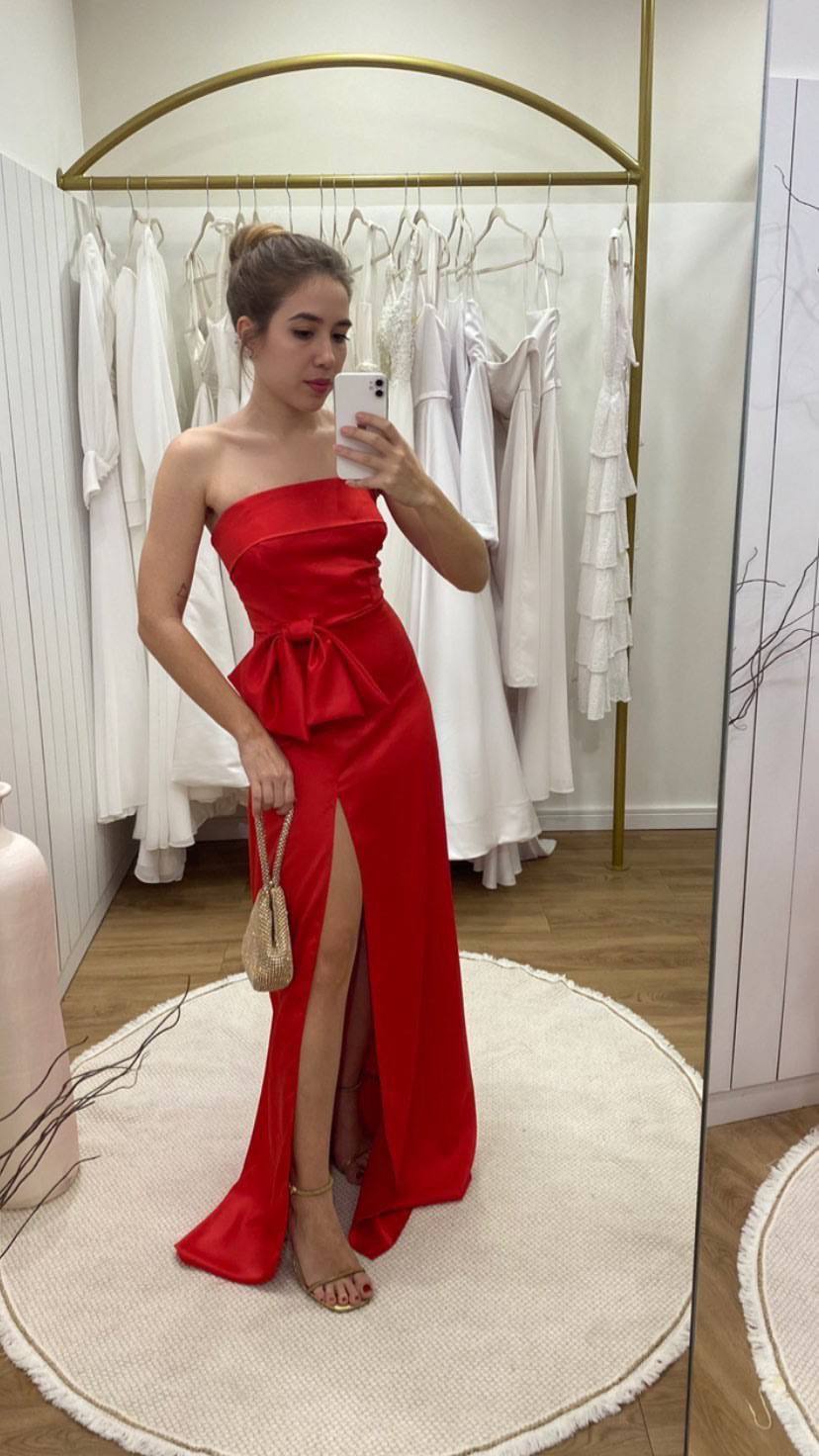 Vestido longo vermelho de cetim, decote tomara que caia, com laço na cintura e fenda frontal pronunciada.