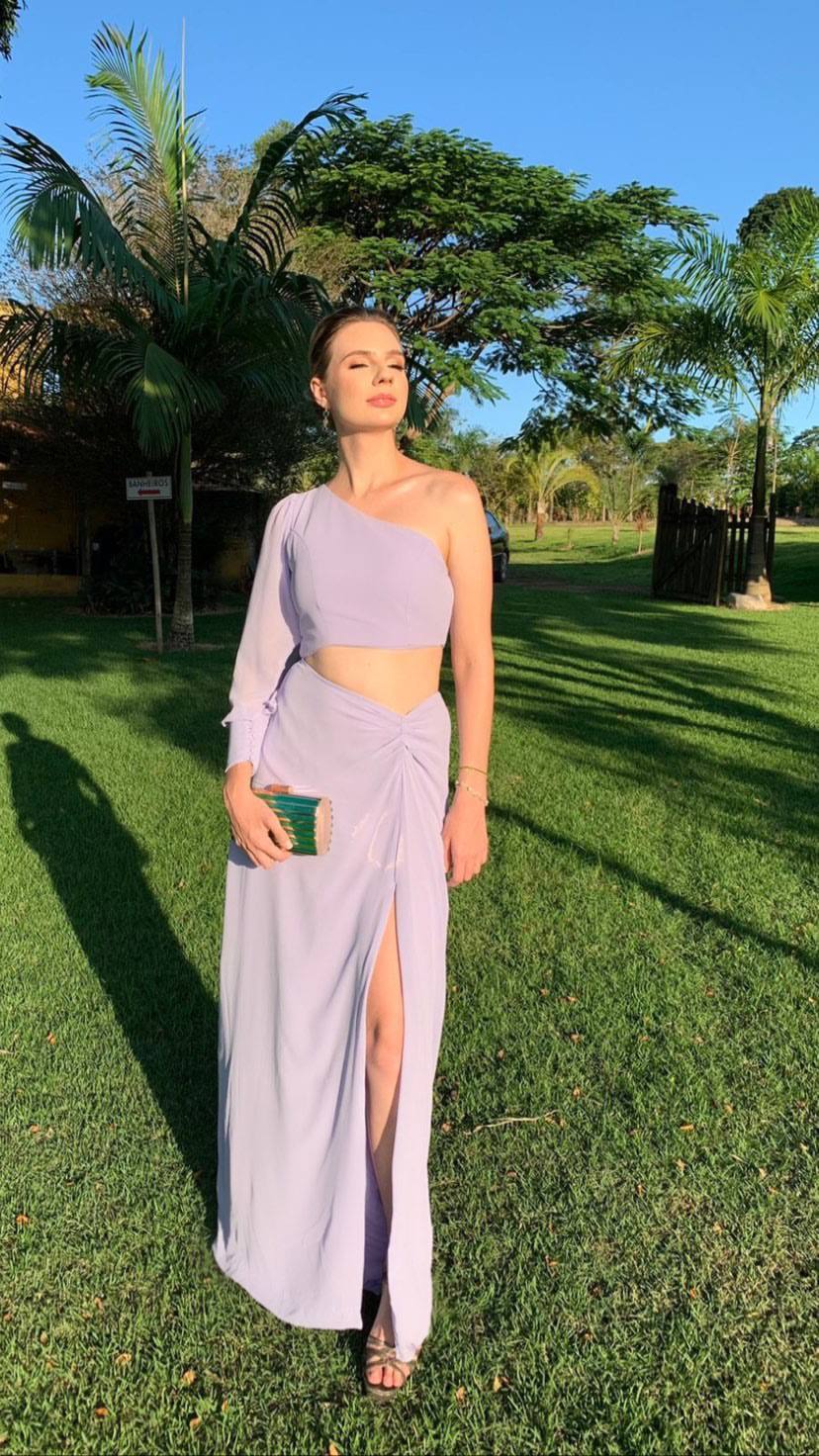 Vestido longo lilás com decote de um ombro, manga longa, recorte na cintura e fenda frontal na saia.