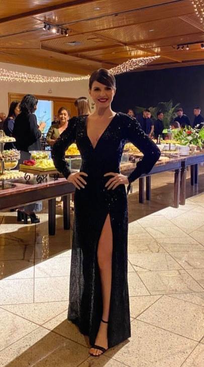 Vestido longo preto com brilho, mangas longas, decote V e fenda frontal alta na saia.