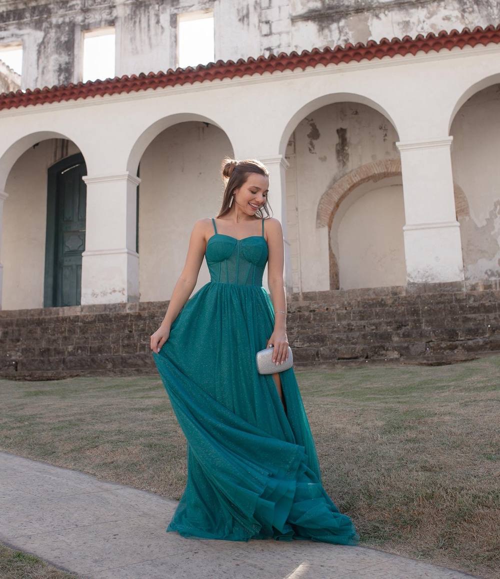 Vestido longo verde petróleo com brilho, de alça fina, corpete estruturado e saia fluida com fenda.